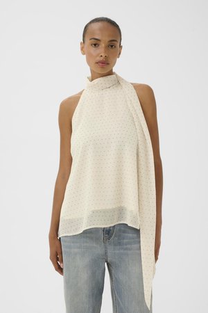 Kvinde iført hvid polkaprik haltertop uden ærmer med stofdragt, kombineret med lyseblå højtaljede jeans, stående mod ensfarvet baggrund.