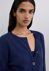 Mujer con cabello oscuro y largo vestida con un cárdigan azul marino con un broche de anillo dorado, en un retrato en primer plano contra un fondo claro y liso.
