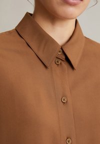 Bruine collared shirt met zachte textuur, voorzien van knoopsluiting en decoratieve knoppen. Eenvoudig, op maat gemaakt ontwerp benadrukt minimalisme.