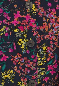 Blommigt tyg med rosa, gula och gröna mönster på en mörk bakgrund, med mörka knappar synliga längs kanten.