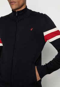 Marinblå zip-tröja med hög krage, med röda och vita randiga detaljer på ärmarna och en liten röd logotyp på bröstet.