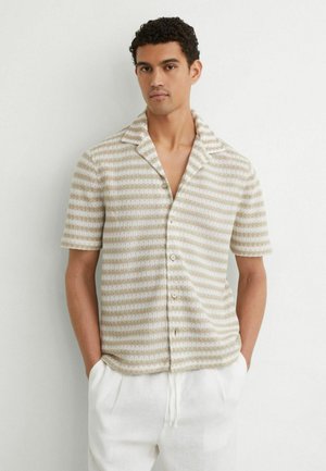 Mand iført kortærmet beige og hvidt, vandret striped strikket shirt med knapper og hvide bukser med snøre, stående mod en hvid væg.