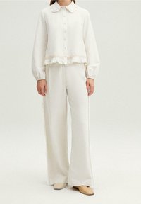 Mujer vistiendo una blusa abotonada color blanco roto con dobladillo con volantes y pantalones de pierna ancha, combinados con unos zapatos planos beige bicolores.