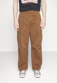 Person trägt braune Cargohosen mit Seitentaschen, ein schwarzes T-Shirt mit "Boogie" Grafik und weiße Sneakers mit Zebra-Streifen.