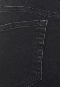 Close-up de tecido de denim preto, destacando um bolso traseiro com costuras decorativas e textura suave, enfatizando a cor escura e as linhas limpas.