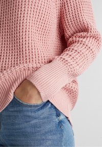  Lichtroze gebreide trui met een textuurpatroon, ribgebreide manchetten en zoom. Gecombineerd met blauwe denim jeans, hand rustend in de zak.