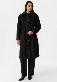 Zwarte wollen mix jas met een kraag, voorknopjes en een riem. Bevat zijzakken en een gestructureerde afwerking. Combinatie met donkere jeans en hakken.