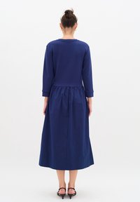 Marinblå klänning med åtsittande topp och hel längd kjol. Långa ärmar, samlad midjedetalj och lättviktig tyg.