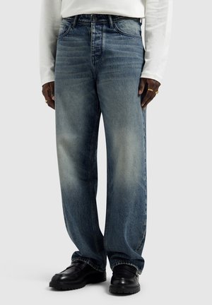 Persoon draagt vervaagde blauwe wijde jeans, zwarte leren schoenen, wit lange mouwen shirt en gouden ringen aan beide handen.