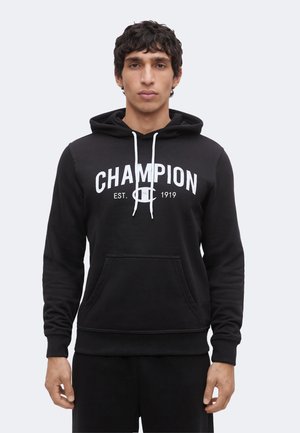 Nuori mies, joka käyttää mustaa Champion-hupparia valkoisilla kiristysnauhoilla ja logotekstillä, seisoo rentoina käsin yksiväristä vaaleaa taustaa vasten.