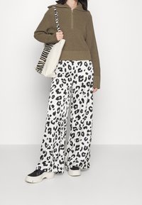 Olivgrüner Strickpullover mit halbem Reißverschluss und weiter schwarz-weiß gemusterter Leopardenhose. Das Modell trägt eine beige Tasche mit einem gestreiften Tragegurt.