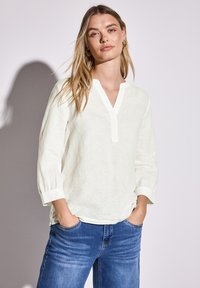 Blouse beige clair avec un col en V, des manches trois-quarts et des détails de bouton. Associée à un jean bleu, doté d'une coupe décontractée et d'un effet délavé.