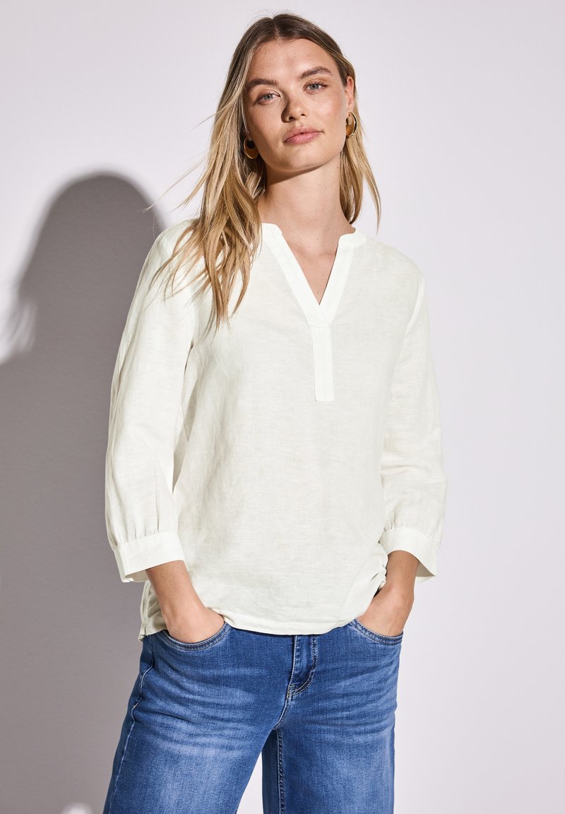 Blouse beige clair avec un col en V, des manches trois-quarts et des détails de bouton. Associée à un jean bleu, doté d'une coupe décontractée et d'un effet délavé.