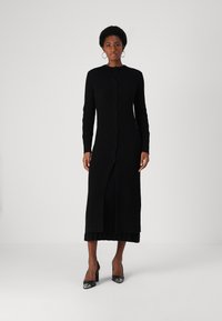 Zwarte geribde gebreide jurk met een lange silhouet, ronde halslijn en een knoopsluiting aan de voorkant. Heeft een zijsplit en lange mouwen.