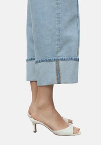 White open-toe mules with a curved heel and soft texture, paired with light blue rolled-up denim pants featuring a contrasting seam.