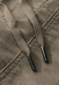 Sweatshirt avec un tissu beige doux et subtil, doté de cordons texturés avec des embouts métalliques et de détails de couture visibles.