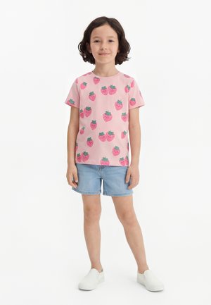 Barn stående iført lyserød t-shirt med jordbærprint, lyseblå denimshorts og hvide slip-on sko på hvid baggrund.