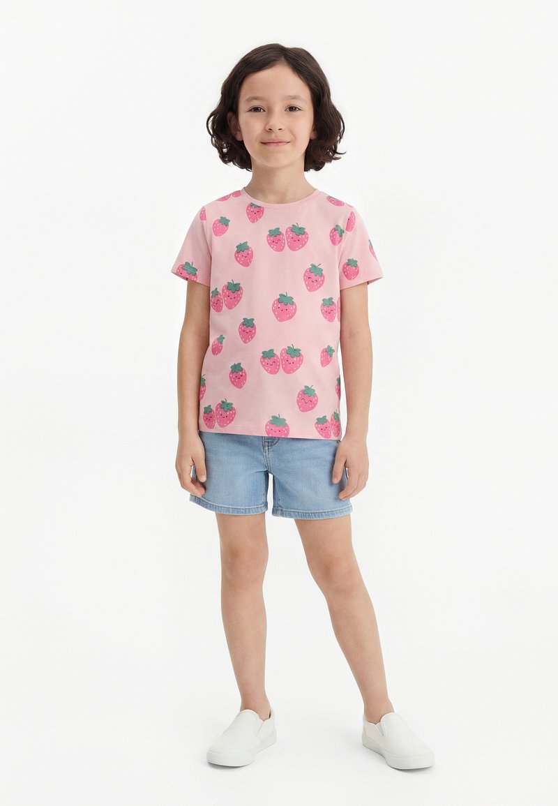 Παιδί που στέκεται, φοράει ροζ μπλουζάκι με κοντά μανίκια και τύπωμα φράουλας, ανοιχτό μπλε τζιν σορτς και άσπρα παπούτσια χωρίς κορδόνια σε λευκό φόντο.