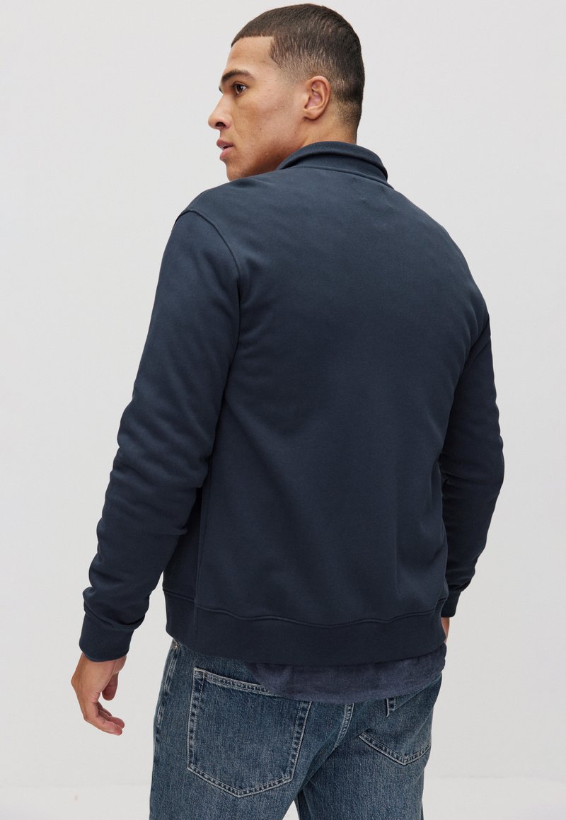 Mörkblå sweatshirt med krage, ribbad ärmslut och nederkant, tillverkad av ett mjukt material. Buren med ljusblå jeans, sedd bakifrån.