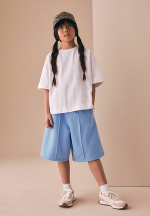 Hosszú fonatokkal rendelkező fiatal, aki leopárdmintás sapkát, fehér, bő méretű pólót, világoskék rövidnadrágot, fehér zoknit és bézs sportcipőt visel.