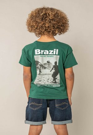 Enfant aux cheveux bouclés portant un t-shirt vert montrant une scène de football en noir et blanc et le texte "Brésil Rio de Janeiro" au dos.