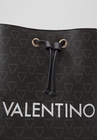 Svart Valentino-väska med textur och vit logotyptext, med en läderdragsko dragen genom öglor i guldfärgad metall.