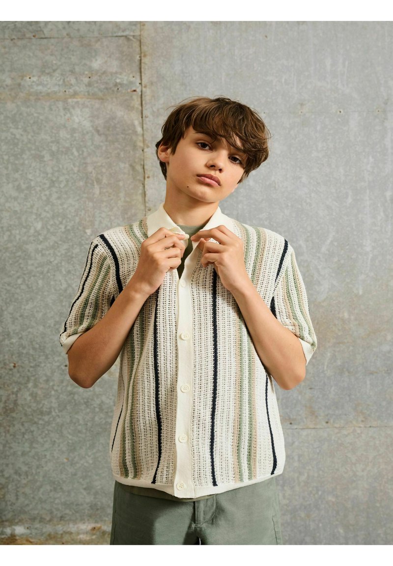 Adolescent garçon ajuste le col d'une chemise en tricot à manches courtes avec des rayures verticales vertes, noires et beige devant un mur texturé gris.