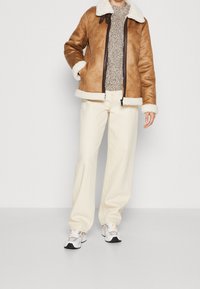Giacca di shearling marrone con rifiniture bianche e cerniera; maglione lavorato a maglia testurizzata; pantaloni a gamba larga beige; sneakers grigie e bianche.