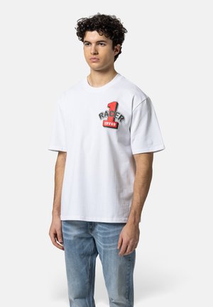 Jonge man met krullend haar, gekleed in een wit oversized T-shirt met rode opdruk "Racer 1 19V69" en lichtblauwe jeans, staande tegen een witte achtergrond.