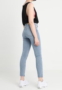 Persona vistiendo jeans ajustados de color azul claro, zapatillas blancas, top negro sin mangas y con un sujetador rosa visible, de pie contra un fondo blanco liso.