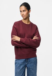 Mulher com cabelo escuro preso, usando brincos de argola, um sweatshirt bordeaux e jeans azuis, de pé com os braços cruzados, olhando para o lado.