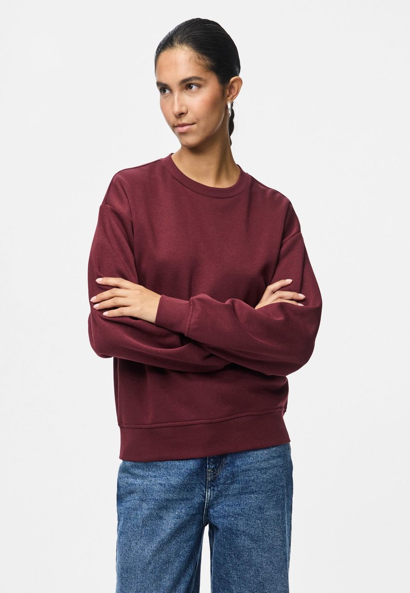 Mulher com cabelo escuro preso, usando brincos de argola, um sweatshirt bordeaux e jeans azuis, de pé com os braços cruzados, olhando para o lado.