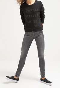 Mustapaita, jossa lukee "SPARKLE GLITTER SHINE" kimaltelevalla tekstillä, yhdistettynä harmaisiin skinny-farkkuihin ja mustiin slip-on-kengiin.