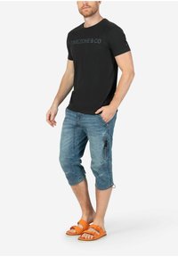 Schwarzes T-Shirt mit "TIMEZONE&CO"-Logo, blaue Jeans-Cropped-Hosen mit Seitentasche und orangefarbene flache Sandalen mit Schnallenakzenten.