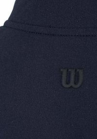 Tejido azul marino con textura suave, que presenta un logo embosado en negro con la letra "W". Las costuras están visibles a lo largo de los bordes del tejido.