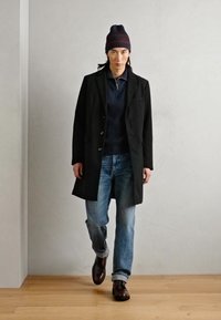 Abrigo negro, suéter azul marino con cremallera, jeans azul claro y zapatos marrones oscuros. El conjunto se complementa con un gorro a rayas azul marino.