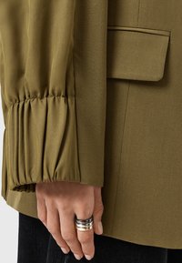 Giacca verde oliva con maniche arricciate e una sola tasca, abbinata a una mano che indossa tre anelli metallici. Texture semplice e liscia.