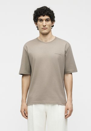 Young man with curly dark hair wearing a taupe oversized short-sleeve t-shirt and white pants, standing against a plain white background.