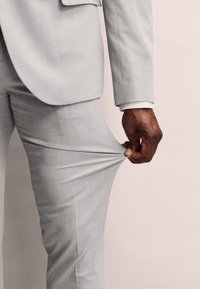 Completo grigio chiaro su misura con un tessuto texturizzato. Una mano tira il gambale dei pantaloni, evidenziando il design aderente e il materiale morbido.