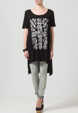Tunique noire à manches courtes avec un imprimé floral gris et blanc, dotée d'un col rond et d'un ourlet arrière plus long, associée à un jean en denim clair.