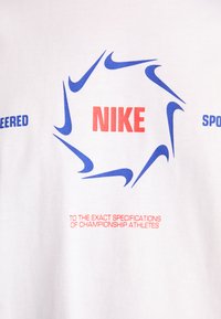 Baltas audinys su keliais mėlynais Nike swoosh logotipais, išdėstytais ratu aplink raudoną "NIKE" tekstą ir mažą raudoną tekstą žemiau.