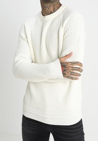 Vaalean kerma, uurrettu neulepusero, jossa on pyöreä kaula-aukko ja pitkät raglanhihat. Täydentää teksturoitu yksityiskohta ja rento mitoitus.