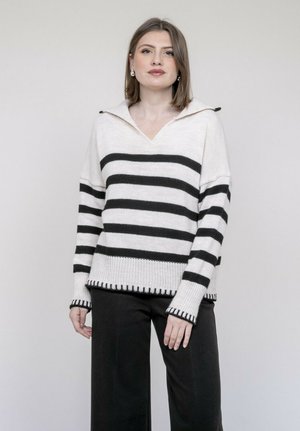 Femme aux cheveux mi-longs portant un pull blanc à rayures horizontales noires et un pantalon noir, debout devant un fond uni.