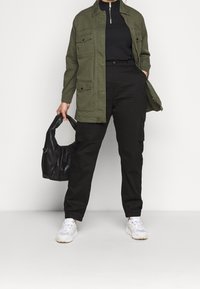 Oliven grøn jakke med flere frontlommer, parret med sorte cargo-bukser og hvide sneakers. Personen holder en sort håndtaske.