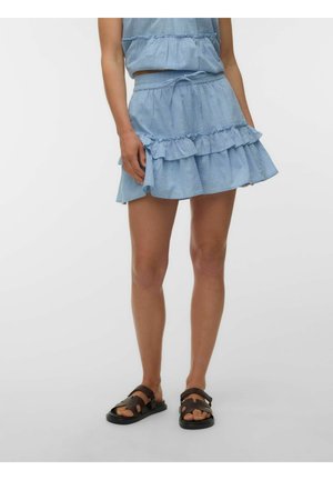 Femme portant une jupe bleue claire à volants avec une taille à cordon et des sandales noires ouvertes à l'avant, debout devant un fond blanc uni.