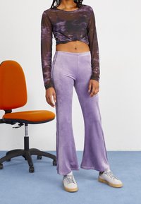 Person som bär en genomskinlig svart och lila långärmad crop-top, lavendelfärgade velourbyxor med vida ben och vita sneakers, står bredvid en orange kontorsstol.