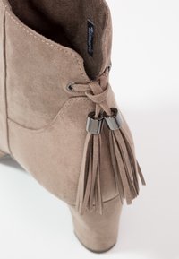 Tanfarbene Wildleder-Ankle-Boots mit hohem Absatz, die ein Schnürdesign und zwei Quasten mit metallischen Akzenten am Knöchel aufweisen. Klassische Steppnähte.
