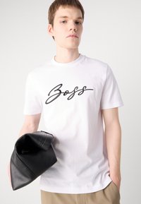 Fehér pamut póló fekete "Boss" hímzéssel, kerek nyakkivágással, rövid ujjakkal. A modellen egy fekete bőr alkalmi táska van.