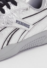 Srebrni športni copati s razpokanimi kovinskimi površinami, črnimi vezalkami, sivim podplatom in blagovno znamko "PLEASURES" na straneh.