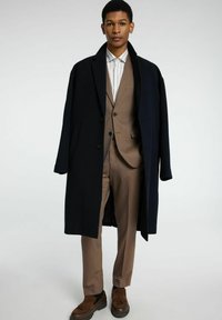 Marine overjas gedragen over een beige pak, met een overhemd met kraag en bruine loafers met kwastdetails. Effen kleuren en een getailleerde pasvorm.
